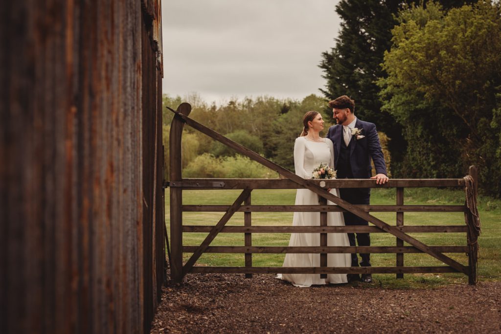 Bride and groom beside wooden gate, countryside background.