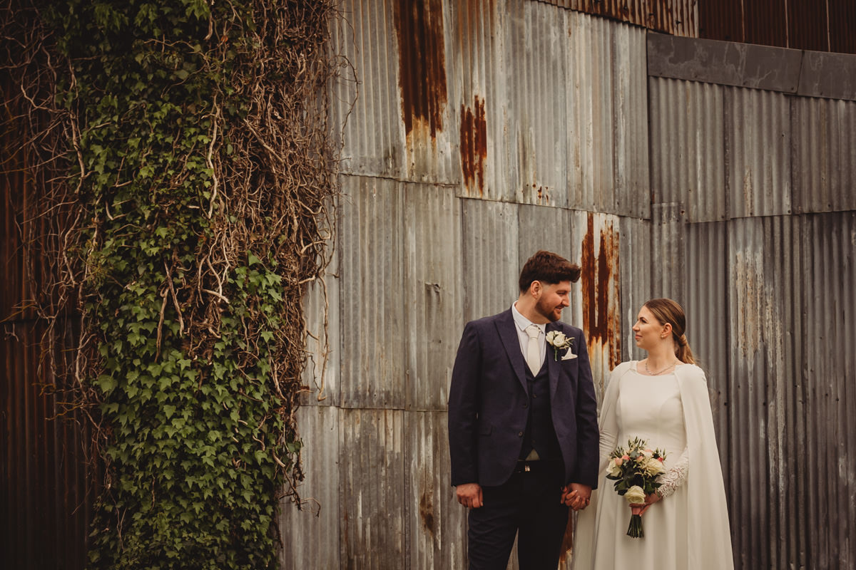 Bride and groom standing by a rustic wall.