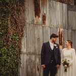 Bride and groom holding hands by rustic wall.