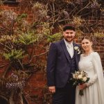 Couple in formal attire by brick wall with flowers.