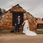 Bride and groom pose with dog by rustic building.