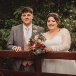 Bride and groom smiling on wooden bridge holding bouquet.