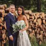 Bride and groom by stacked logs in forest.