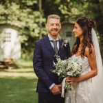 Bride and groom smiling outdoors, holding flowers.