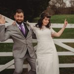 Couple posing playfully by a white gate outdoors.