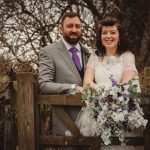 Bride and groom smiling with flowers by wooden gate.