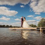 Couple posing on lakeside pier under blue sky.