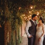 Wedding couple smiling under string lights and greenery.