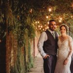 Couple smiling under leafy arch with lights.