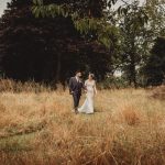 Bride and groom walking in a grassy field.