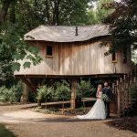 Couple poses by rustic treehouse in wooded area.