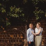 Couple posing against brick wall with tree backdrop.