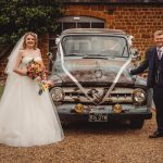 Bride and groom pose by vintage wedding car.