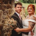 Smiling couple in wedding attire with tattoos and bouquet.