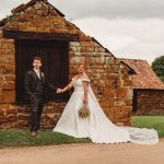 Bride and groom standing by rustic stone building.