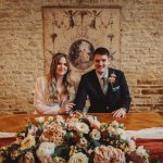 Wedding couple seated at decorated table, rustic backdrop.