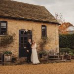 Couple standing outside rustic stone building, autumn garden.