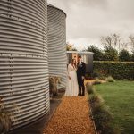 Couple standing beside rustic silos and garden path.