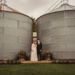 Wedding couple between large metal silos outdoors.