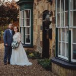 Couple in wedding attire outside stone house.