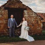 Bride and groom holding hands by rustic building.