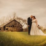 Couple embraces in field near rustic wooden barn.