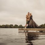 Couple standing on lakeside pier, cloudy sky.