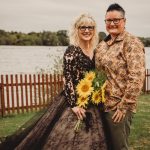 Couple posing with sunflowers by a lakeside fence.