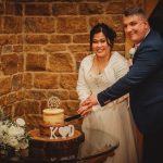 Couple cutting wedding cake beside rustic wall.