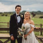 Outdoor wedding couple smiling by countryside fence.