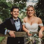 Smiling couple in wedding attire by wooden fence.