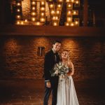 Bride and groom under 'LOVE' sign, rustic venue.