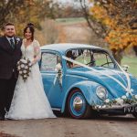 Newlyweds pose beside a vintage blue car.