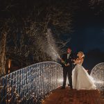 Newlyweds celebrate on a lit bridge with champagne.