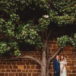 Wedding couple under a large tree by brick wall.