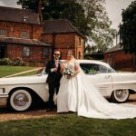 Bride and groom pose by vintage car outdoors.