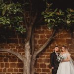 Bride and groom standing by brick wall and tree.