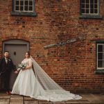 Bride and groom standing by brick cottage wall.