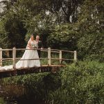 Bride and groom on rustic bridge in green landscape.