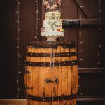 Rustic wedding cake on wooden barrel with lights.