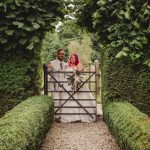 Bride and groom at garden gate, lush greenery.