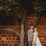 Bride and groom pose under tree against brick wall.