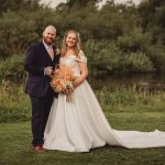 Smiling couple in wedding attire by a river.