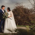 Bride and groom smiling outdoors, bouquet in hand.