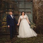 Bride and groom holding hands outdoors, rustic backdrop.