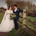 Bride and groom leaning on wooden fence outdoors.