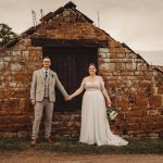 Bride and groom holding hands by rustic stone building.