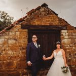 Bride and groom holding hands near rustic stone building.