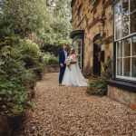 Bride and groom in garden pathway by stone building.