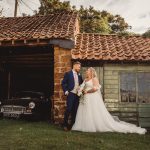 Bride and groom beside rustic garage and vintage car.